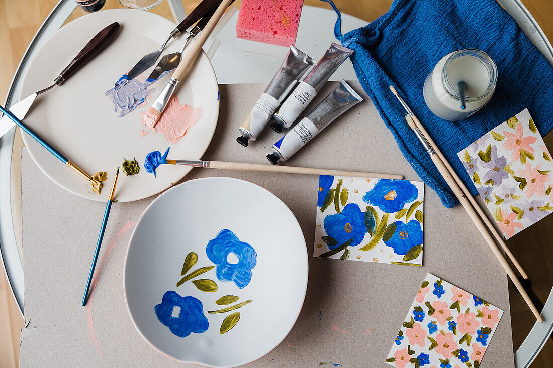 Painted floral ornament on white plate placed on table with set of paint tubes and paintbrushes