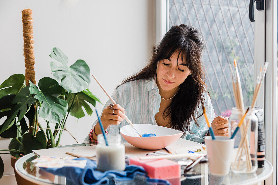 Concentrated talented female artist painting ceramic bowl while sitting at table with set of paintbrushes
