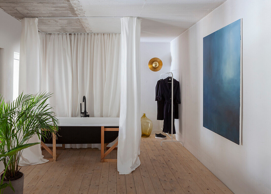 Freestanding bathtub behind curtains in minimalist room with wooden floor