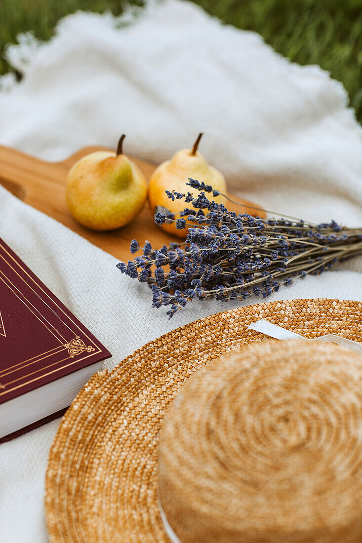 Holzbrett mit Birnen, Lavendelblüte, Strohhut und Buch auf Picknickdecke