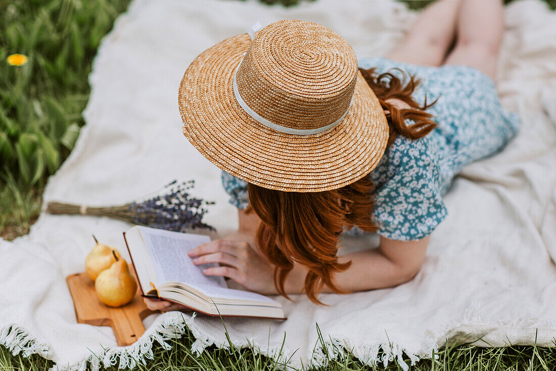Rothaarige Frau im Kleid und Strohhut liegt auf einer Picknickdecke und liest