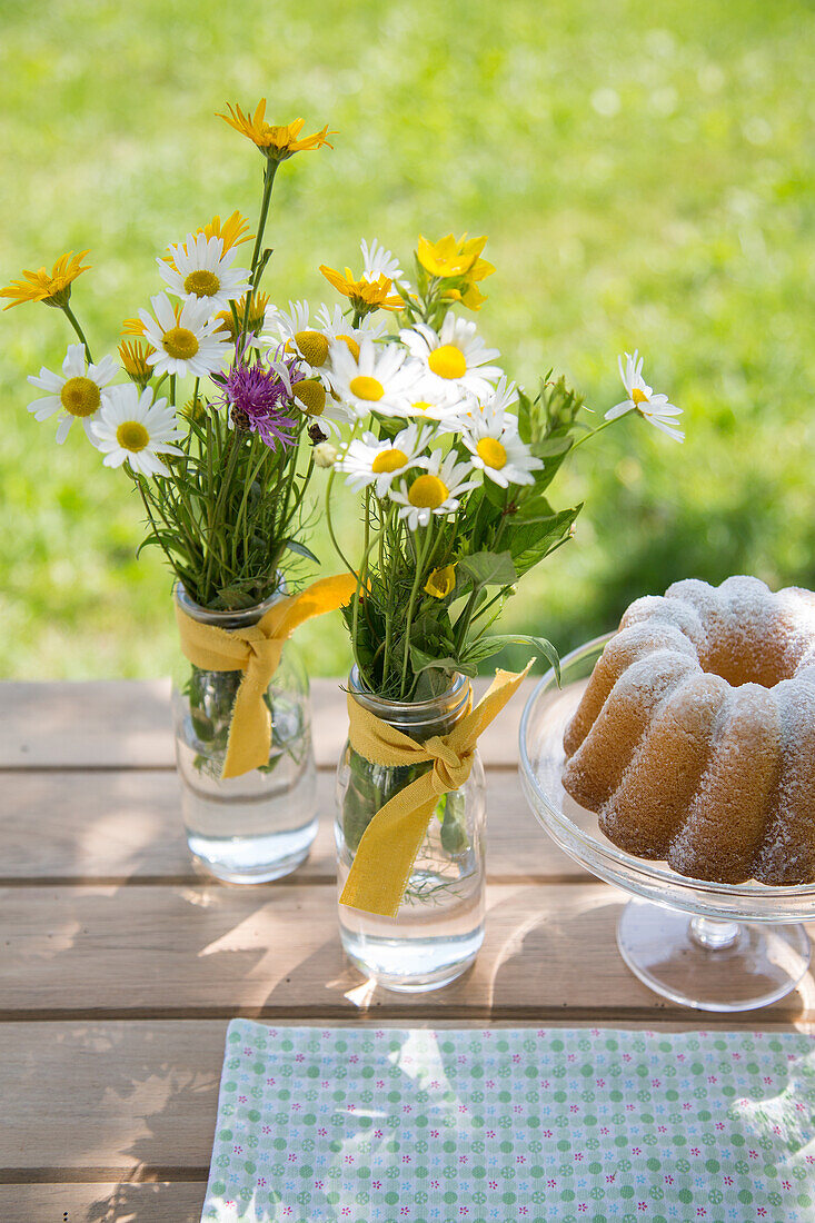 Wildblumensträuße in Glasvasen und Gugelhupf auf Holztisch im Garten