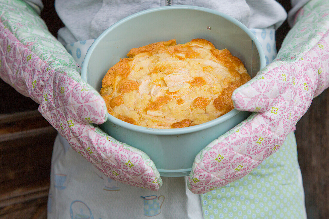 Freshly baked cake in cake tin, held by hands in oven gloves