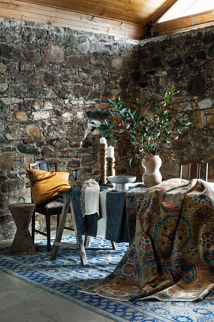 Table with various textiles, vase with leafy branches, candle holder and chair in rustic room with natural stone walls