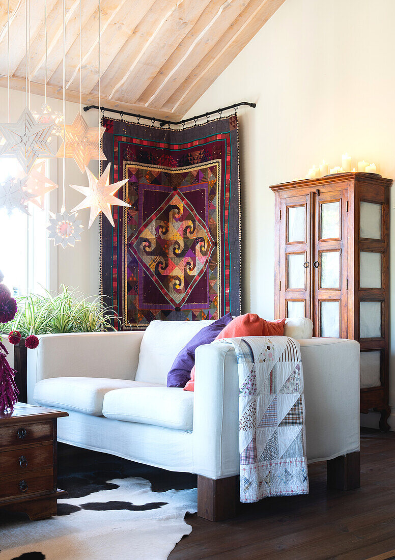 Light-colored sofa in front of cupboard and tapestry, white-painted wooden Advent stars hanging from the ceiling