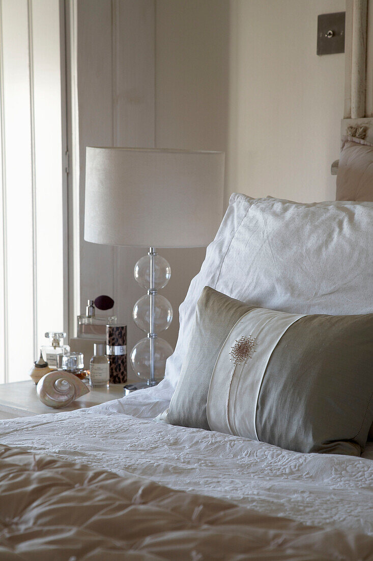 Lampenschirm und Parfümflaschen auf dem Nachttisch mit Kissen und Bettdecke