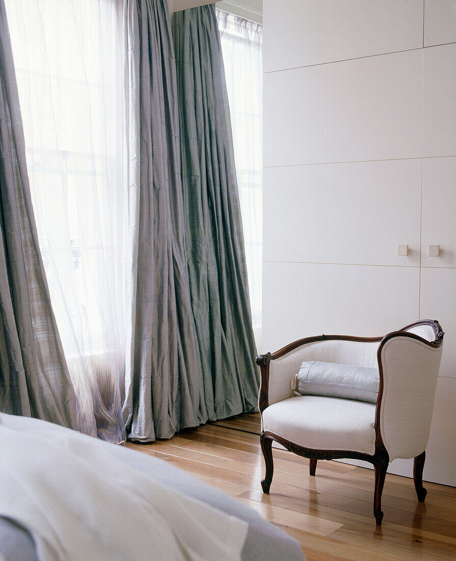 Ein traditionelles Schlafzimmer mit grauen und neutralen Farben, Einbauschränken, Vorhängen und einem gepolsterten Stuhl mit Holzboden