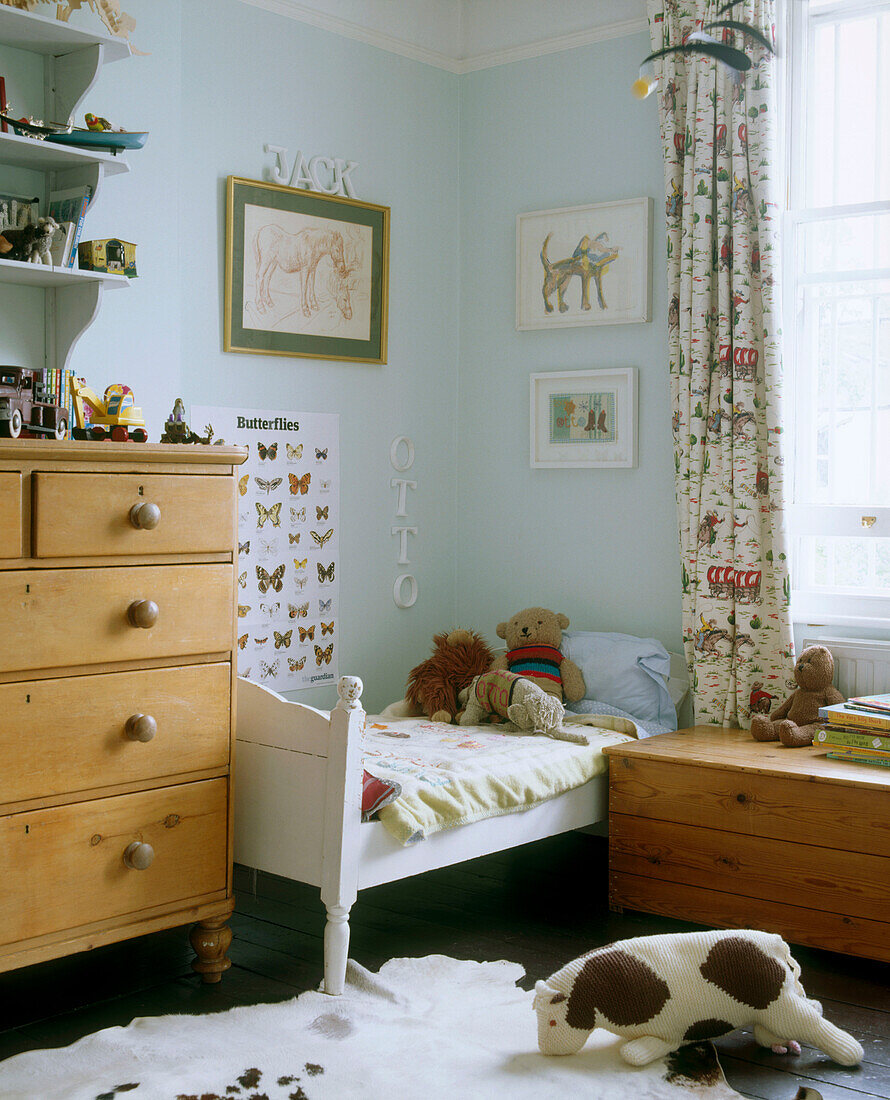 A traditional children's bedroom containing a set of pine drawers next to a small bed surrounded by framed pictures and posters with a wooden chest underneath the window