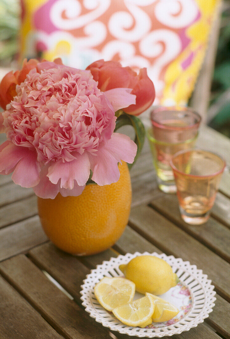 Außendetail einer Vase mit rosa Blumen neben Teller mit halbierten Zitronen auf Holz-Gartentisch