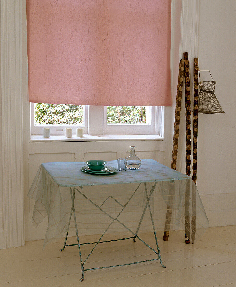 Organzatuch auf dem Tisch mit Geschirr am Fenster mit geschlossenem rosa Rollo