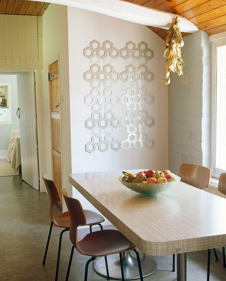 Ein modernes Esszimmer mit einer Glasschale mit Obst auf einem Retro-Esstisch und Stühlen mit einer dekorativen Wanddekoration aus Metall