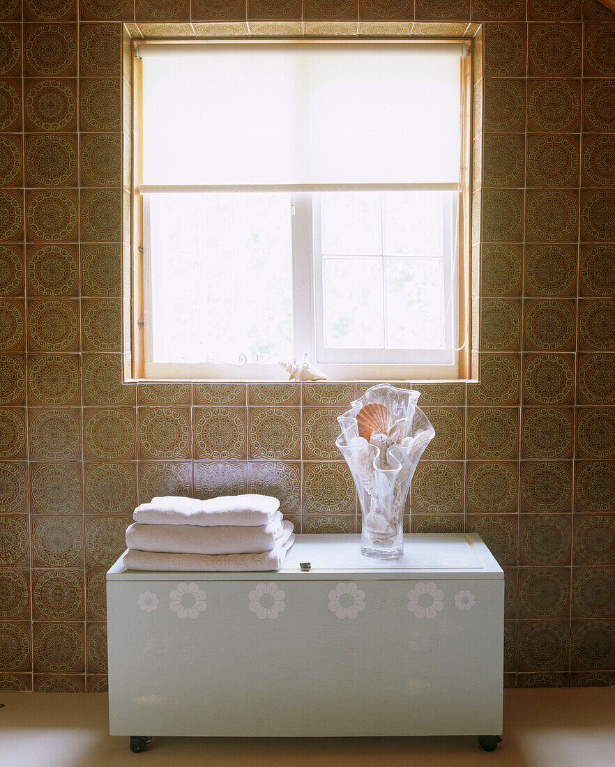 Modernes Badezimmer Details eines Stapels von Handtüchern und einer Glasvase voller Muscheln auf einer dekorierten Badezimmerkommode quadratisches Fenster Retro-Muster Fliesen