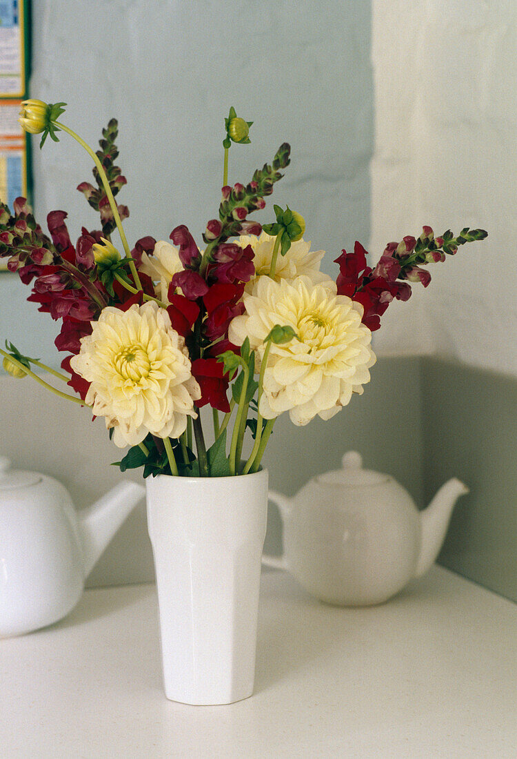 A detail of a white vase of flowers on a table with small white teapots in the background