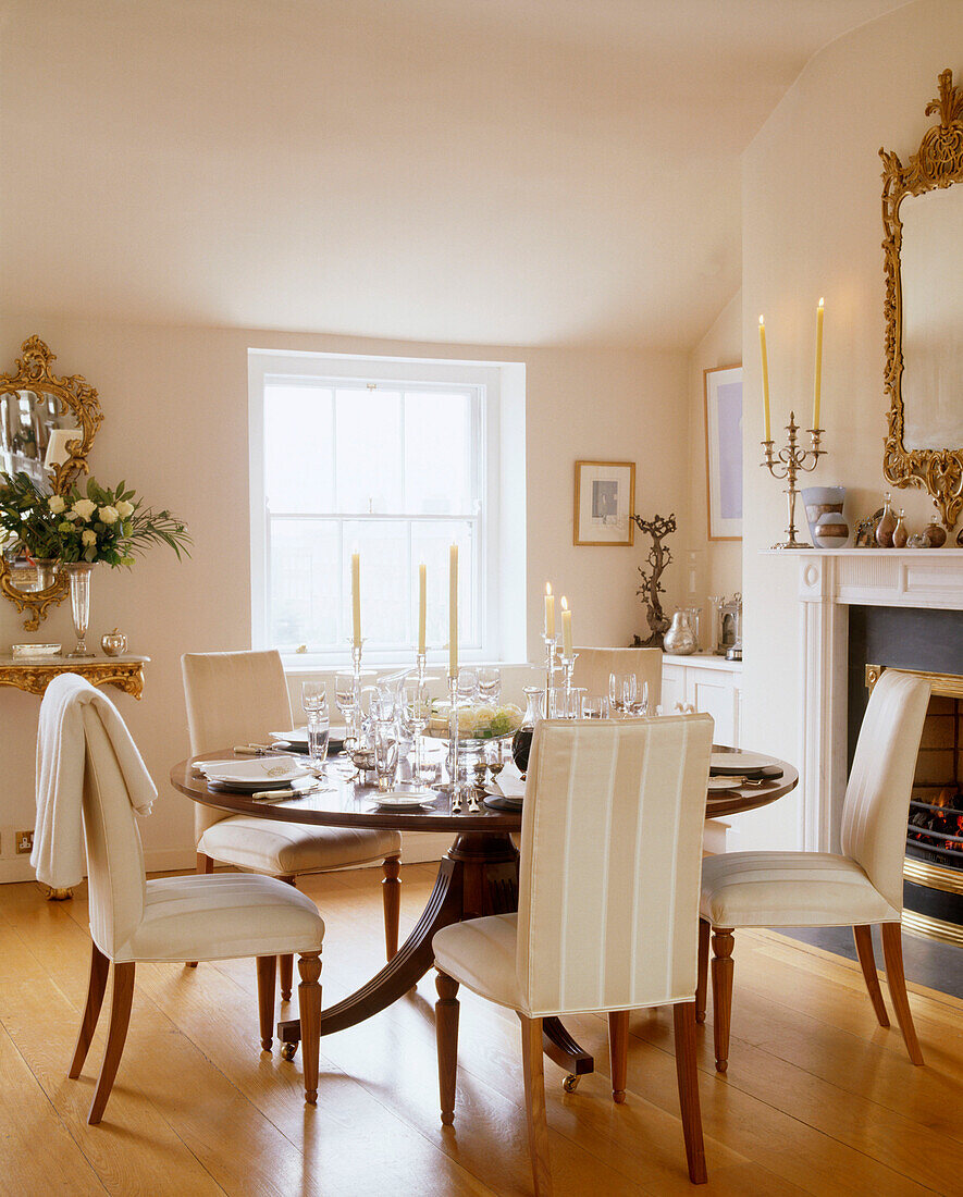 A traditional dining room with a wooden floor and upholstered chairs surrounded a circular table with full place settings and glass candleholders