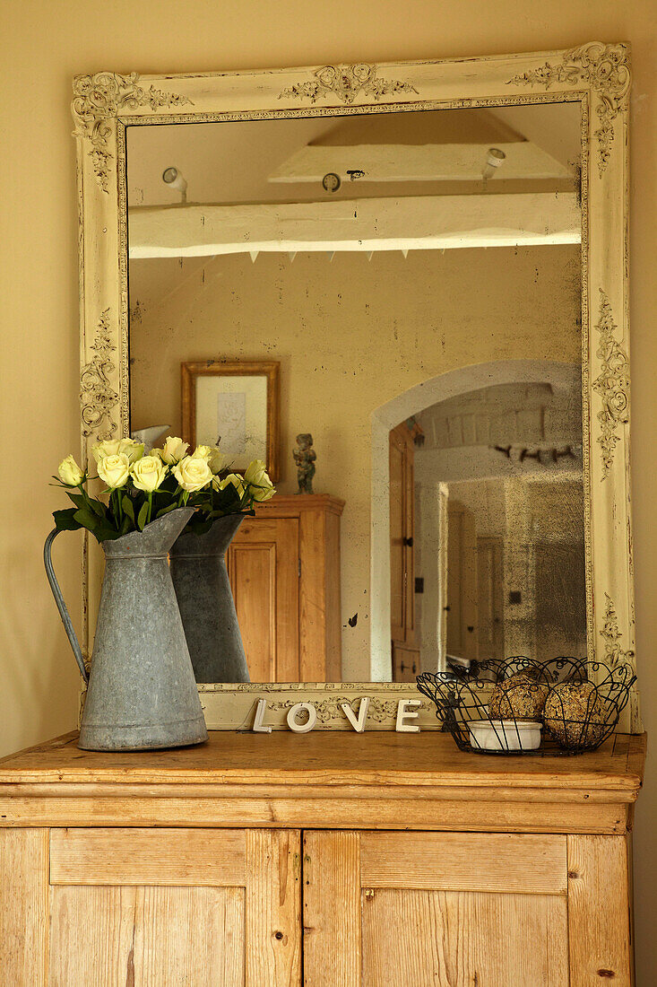 Vintage mirror and metal jug of cut roses on wooden sideboard in West Sussex home, England, UK