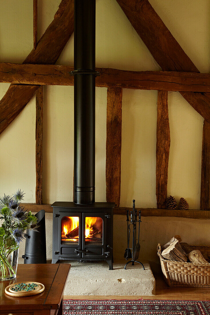 Brennender Holzofen und Korb mit Holzscheiten in einem Haus in West Sussex, England, UK