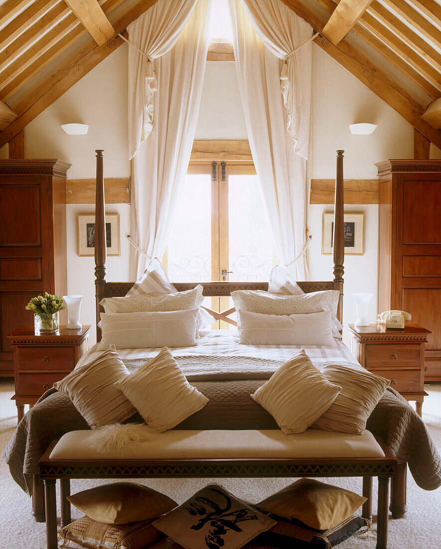 Ein traditionelles holzgerahmtes Doppelbett und eine Bank mit Kissen in einem Schlafzimmer im Landhausstil mit Holzbalken an der Decke einer umgebauten Scheune