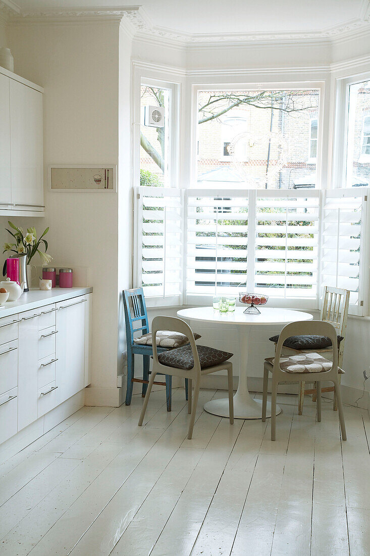 A modern kitchen with a white painted floor and a round kitchen table underneath a large bay window with shutters