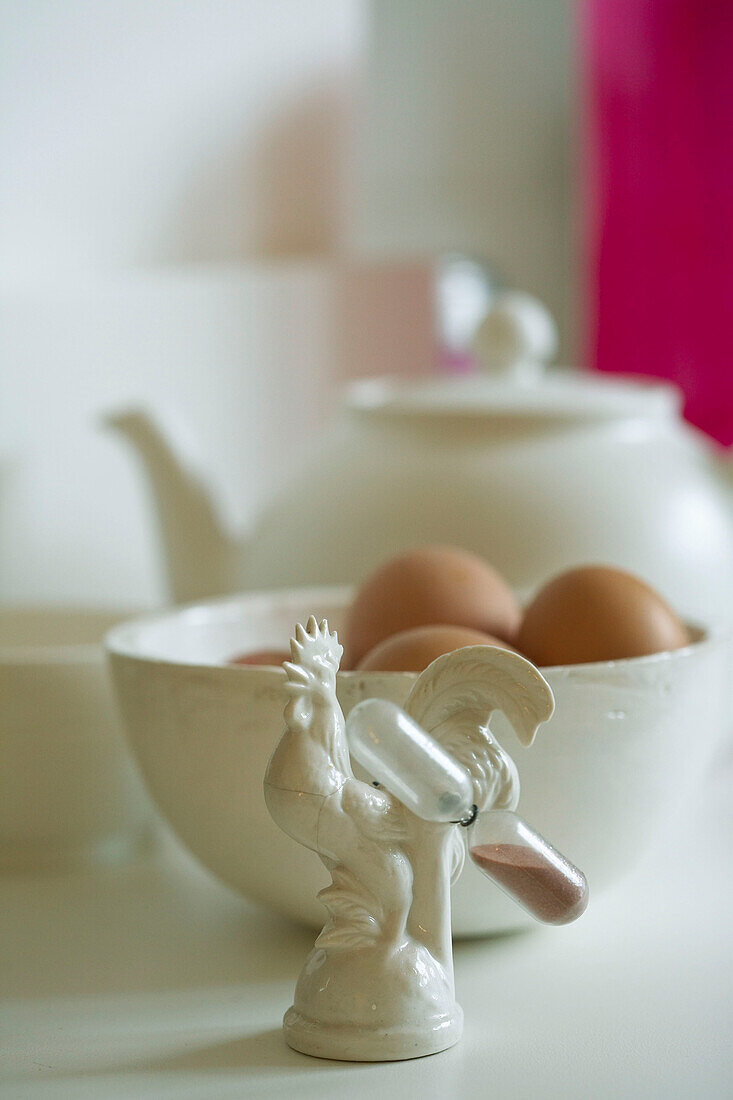 Nahaufnahme einer verschnörkelten Eieruhr dahinter Schale mit Eiern und eine Teekanne