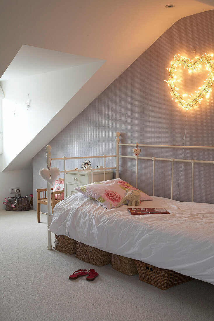 A modern child's bedroom with a large sloping ceiling and fairy lights in the shape of a heart above the traditional iron cast bed