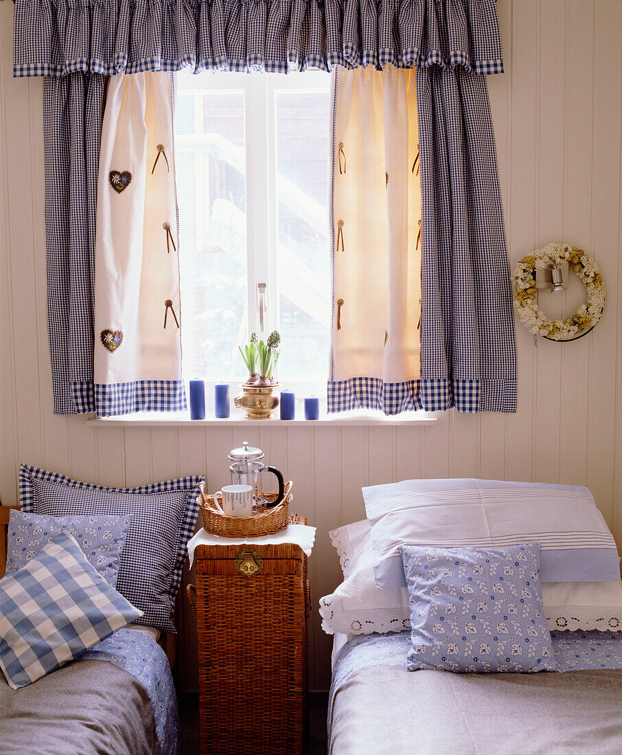 Schlafzimmer im Landhausstil mit zwei Einzelbetten, blauen Vorhängen, passender Bettwäsche, Nachttisch aus Weidengeflecht und Tablett mit Kaffeekanne