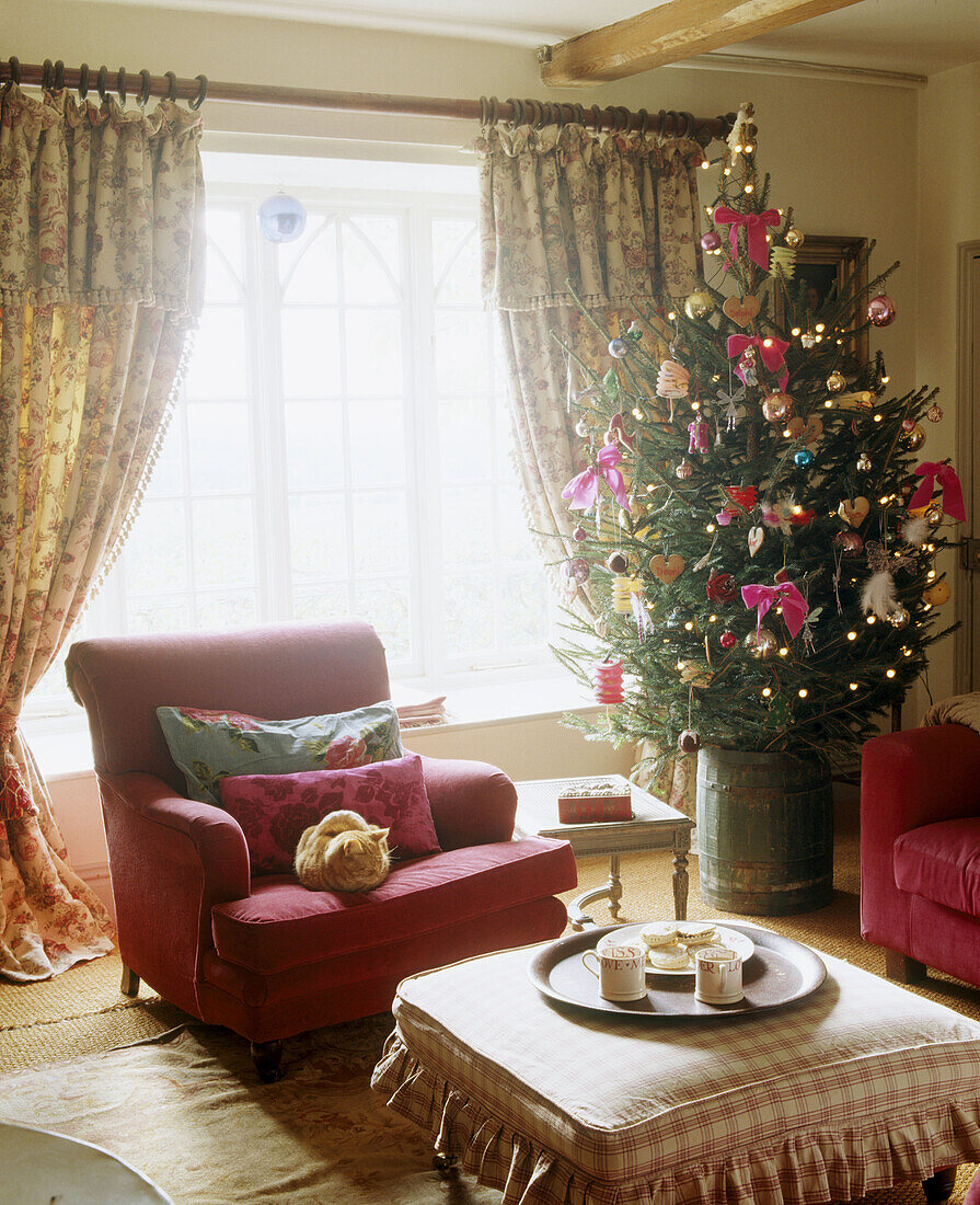 Weihnachtlich geschmücktes traditionelles Wohnzimmer im Landhausstil mit Balkendecke, Weihnachtsbaum, Polstersessel, Ottomane und Vorhängen mit Blumenmuster