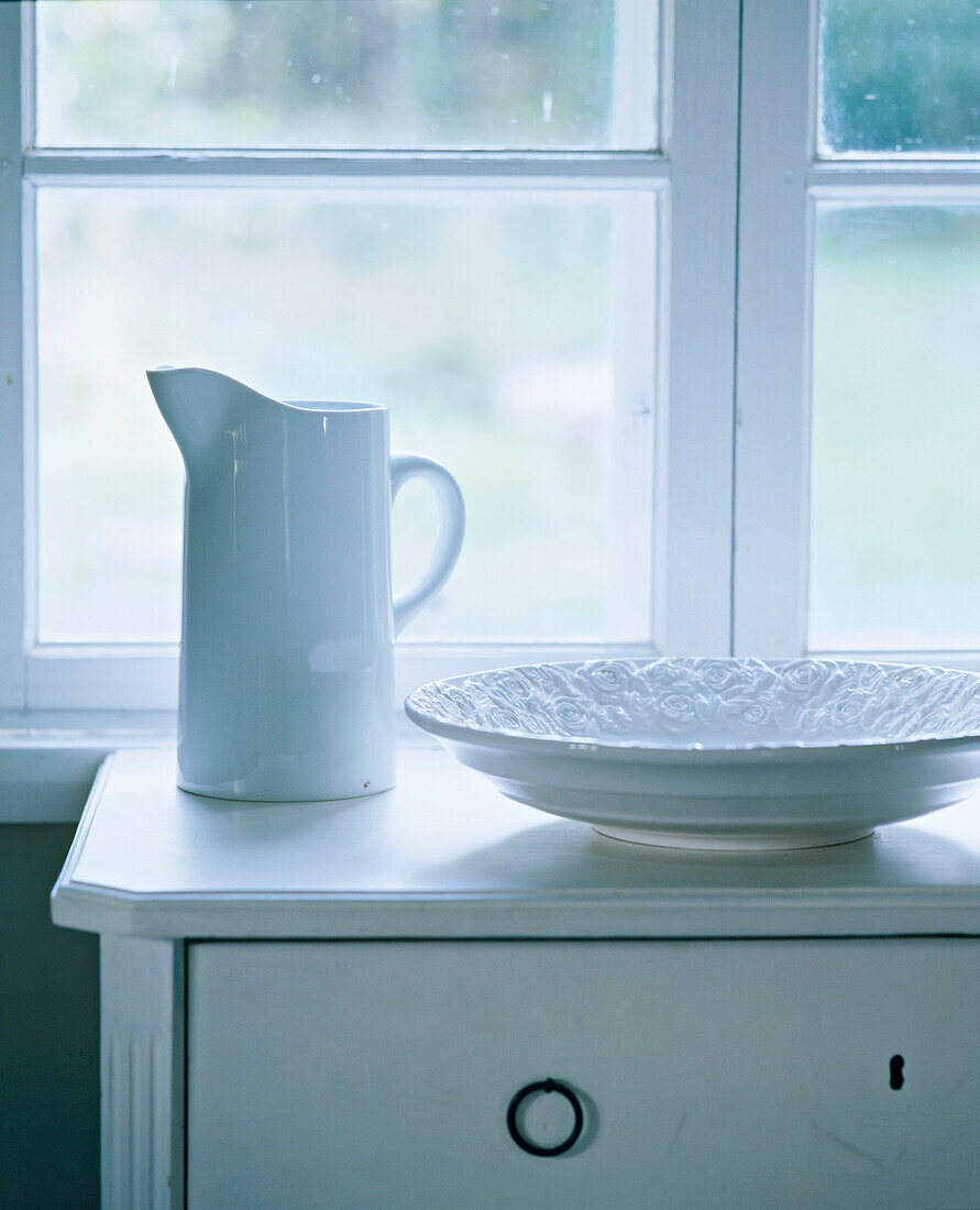 Kommode im Landhausstil mit Keramikkrug und Teller vor einem Fenster