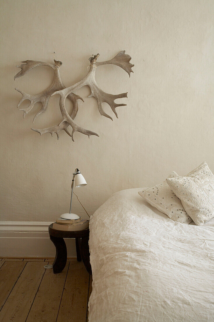 Animal horns on wall above wooden bedside table next to simple double bed with neutral cover