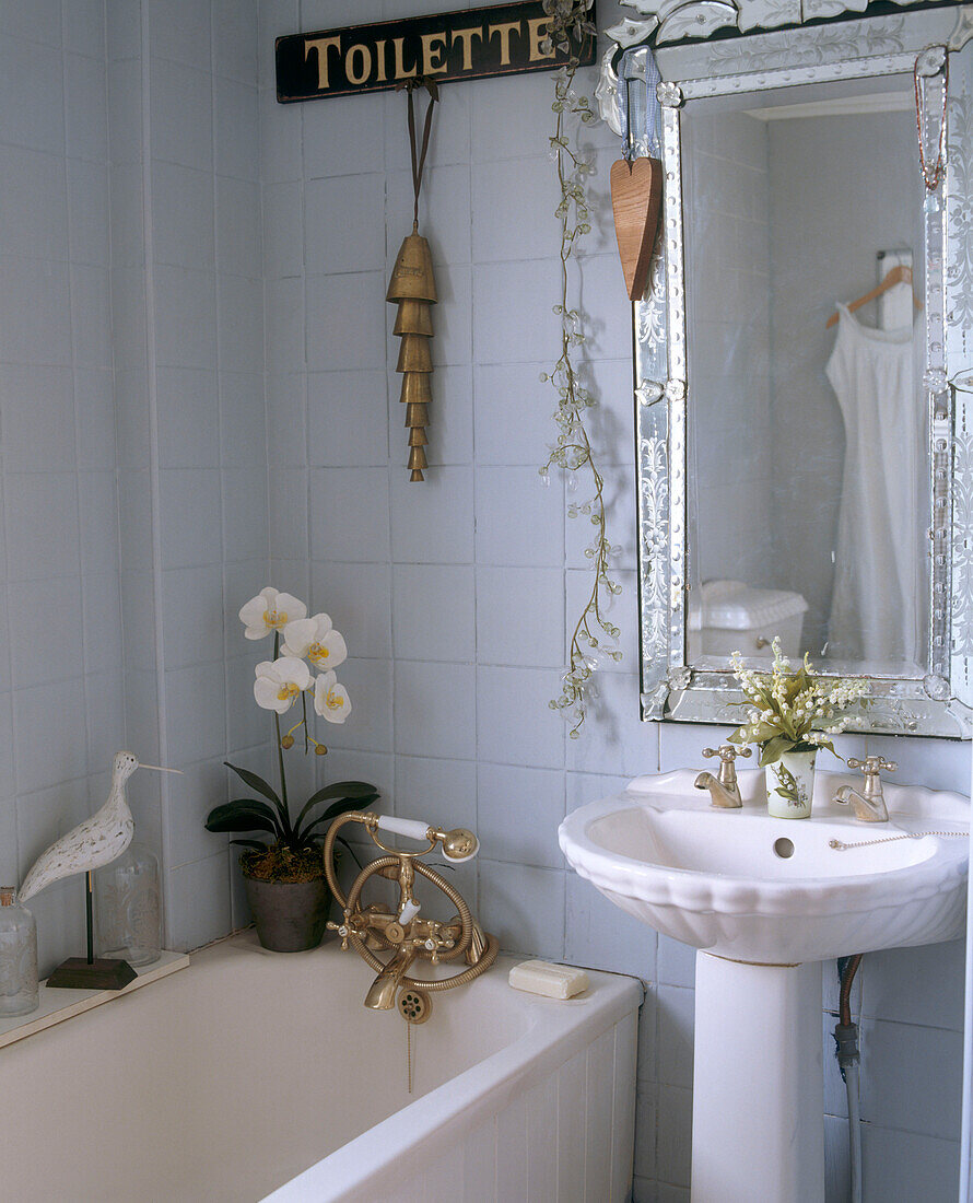 A detail of traditional bathroom with a large silver framed mirror above a sink and next to a bath with a gold taps and shower controls
