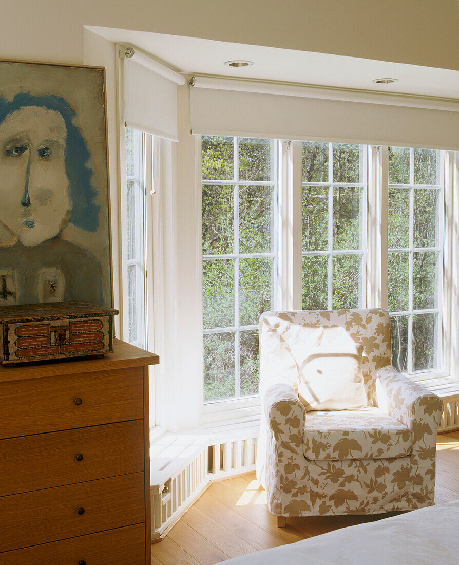 Armchair bathed in light in a bay window area next to a frame painting above a wooden chest of drawers
