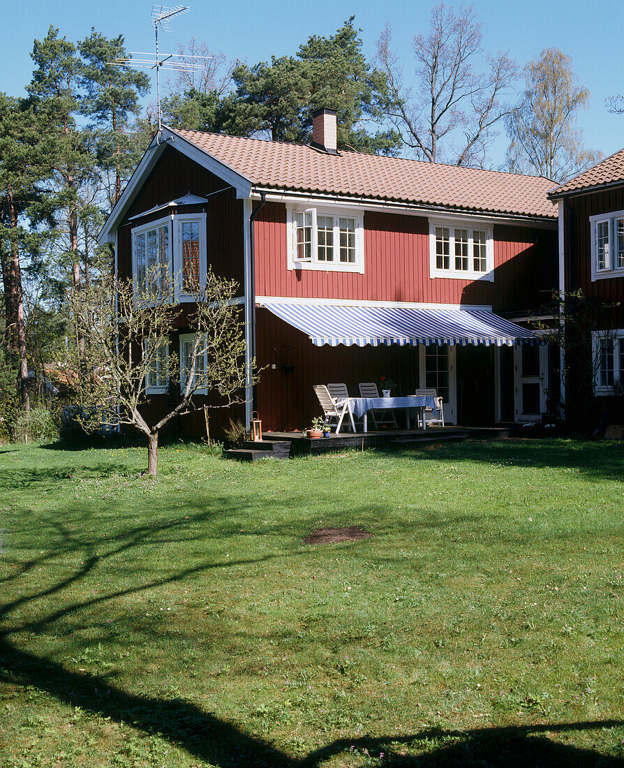 Außenansicht der Rückseite eines traditionellen Holzhauses mit einer Markise über einem Tisch und Stühlen mit Blick auf eine große Rasenfläche