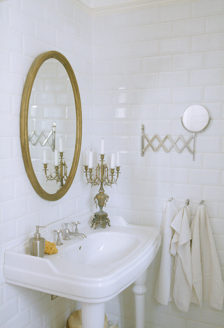 A detail of a traditional bathroom wash basin below a circular gold framed mirror and next to a small candelabra on the edge of the sink