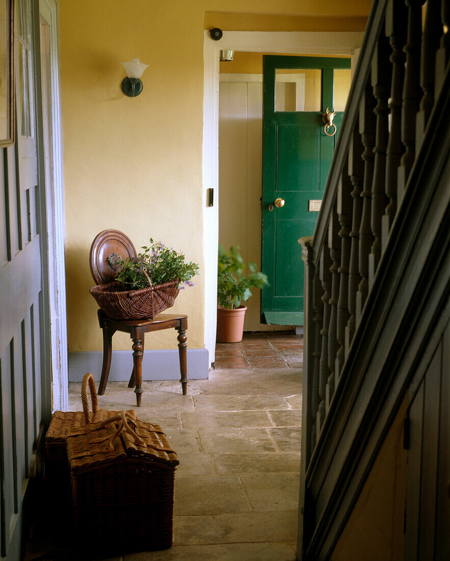 Gelber Flur im Landhausstil mit gefliestem Boden, Treppe, Holzstuhl, Blumenkörben und geöffnter Eingangstür