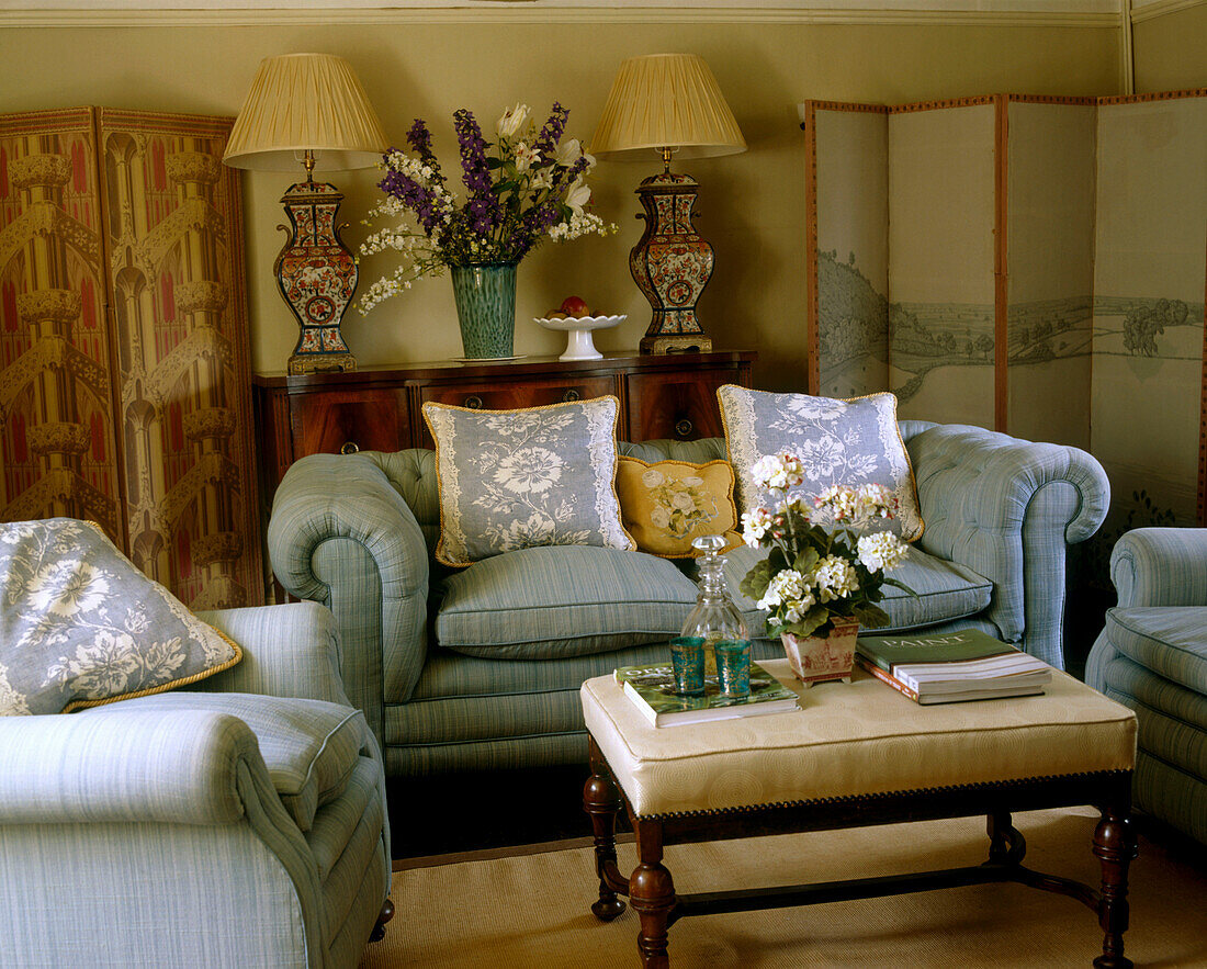 Ein traditionelles Wohnzimmer im Landhausstil mit gepolstertem grünen Sofa, Sessel, Ottomane, Kissen, Tischleuchten und Paravents
