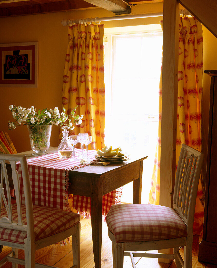 Esszimmer im Landhausstil mit gemusterten Vorhängen, Stühlen mit rotkarierten Polstern, Blumenarrangement, Karaffe und Gläser auf Esstisch aus Holz