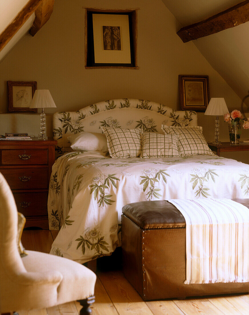 Ein traditionelles Schlafzimmer im Dachgeschoss mit Balkendecke, Doppelbett mit geblümter Bettdecke, Lederhocker und Holzfußboden