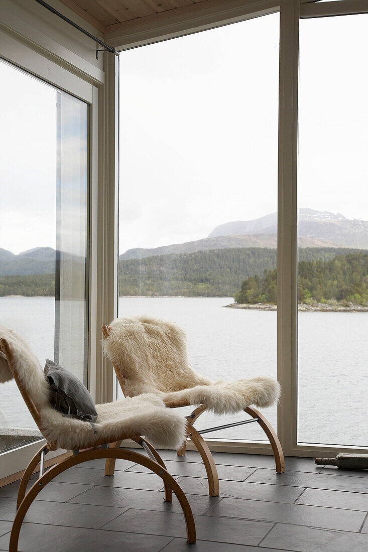 Zwei Holzsessel mit weiße Fellen am Fenster mit Blick auf einen See