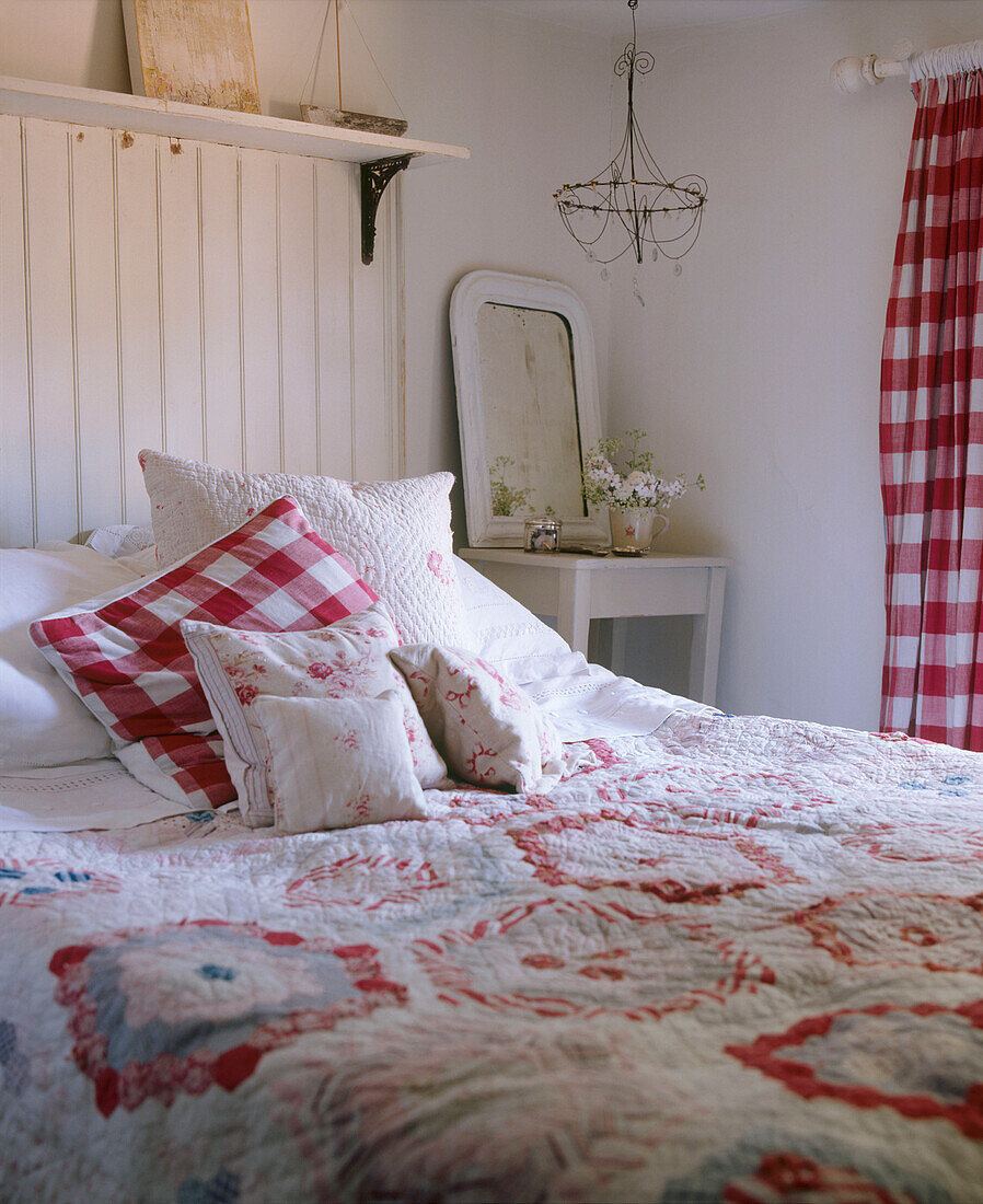 Ein Schlafzimmer im Landhausstil mit roter und weißer Holzvertäfelung, Doppelbett, Patchwork-Steppdecke, Kissen und Vorhängen aus kariertem Stoff, Schminktisch