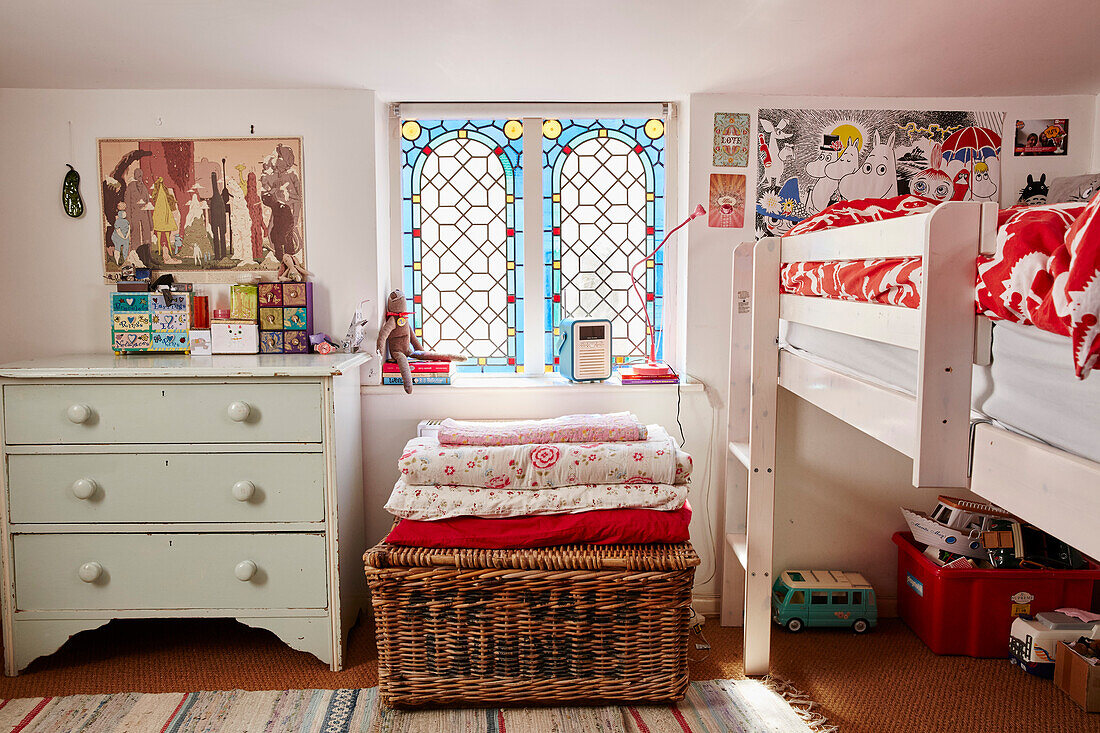 Platform bed with folded bedding on basket below stained glass window in Bridport home, Dorset, UK