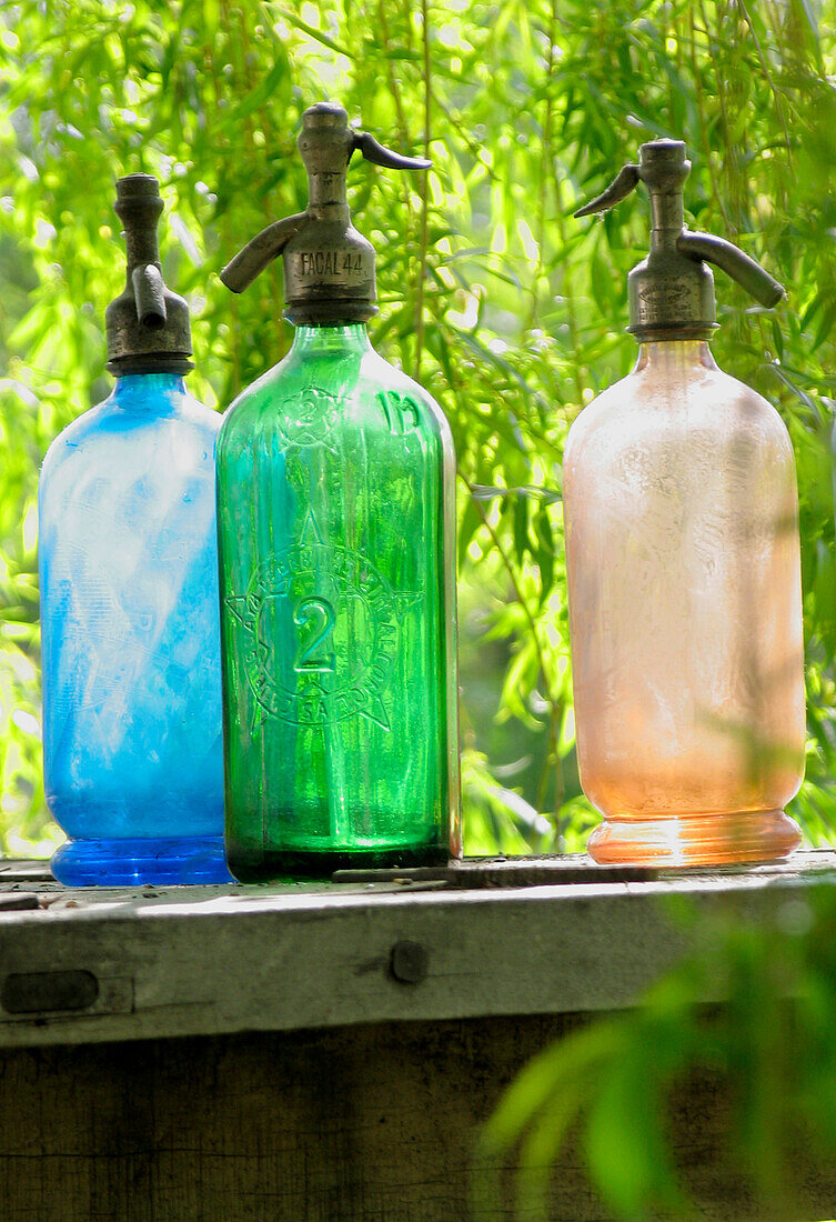 Bunte Soda-Siphons auf einem Gartentisch aus Holz in einem Garten mit leuchtend grünem Blättern