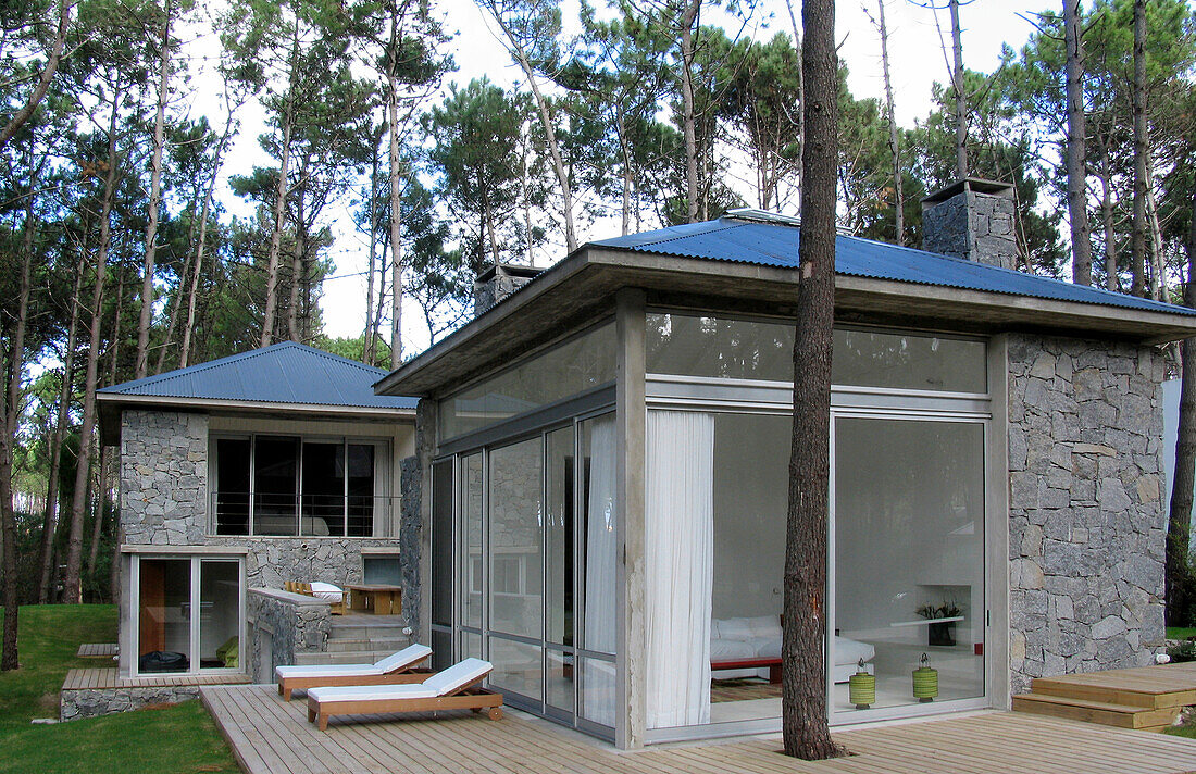 Modernes Haus aus Glas, Holz und Stein mit grauem Wellzinkdach und Holzterrasse in einem Waldgebiet