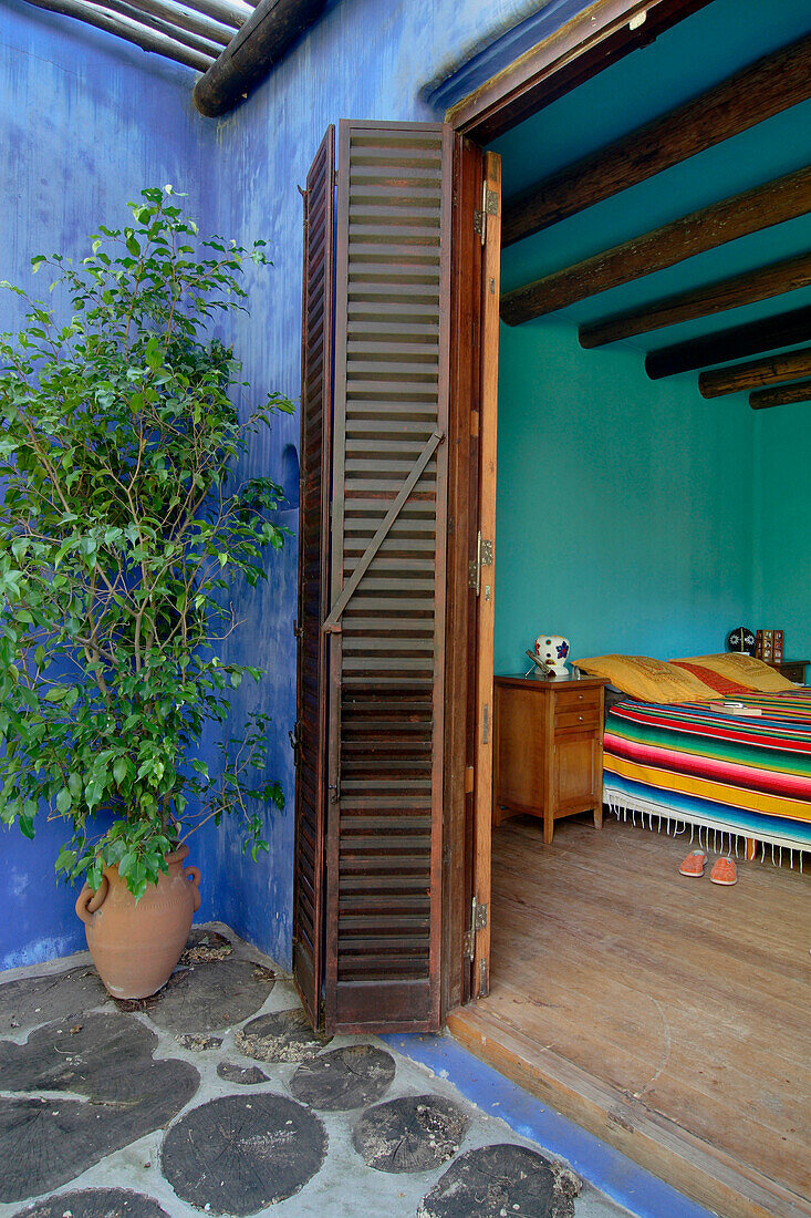 Aquamarinblaues Schlafzimmer mit Balkendecke und mexikanischer Decke auf dem Bett mit Blick durch eine verschlossene Türöffnung