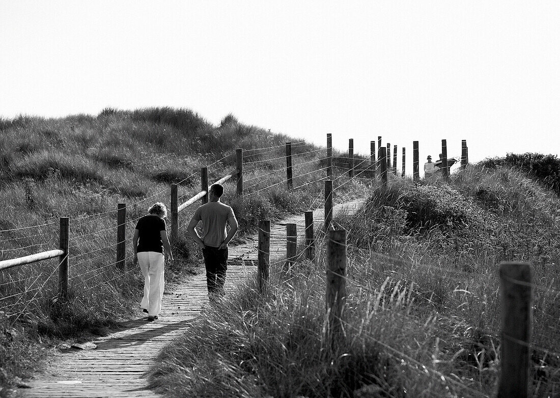 Rückansicht eines Paares, das auf einem Steg in den Sanddünen vor dem Strand spazieren geht