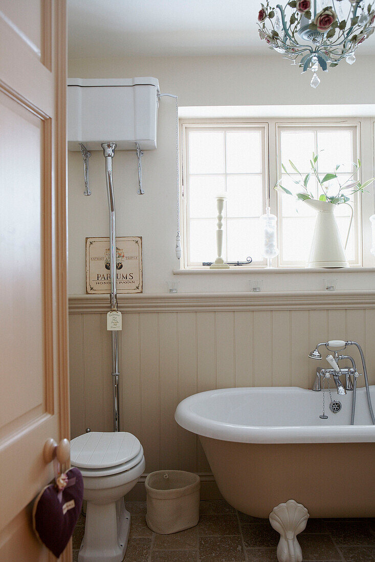 Roll top bath below sunlit window in panelled bathroom with wall mounted cistern
