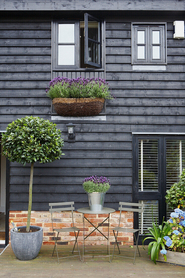 Blick auf die Fassade des Hauses mit schwarzer Klappe, Gartenmöbeln aus Metall und einem Eimer mit Lavendel sowie einem mit Lavendel gefüllten Blumenkasten