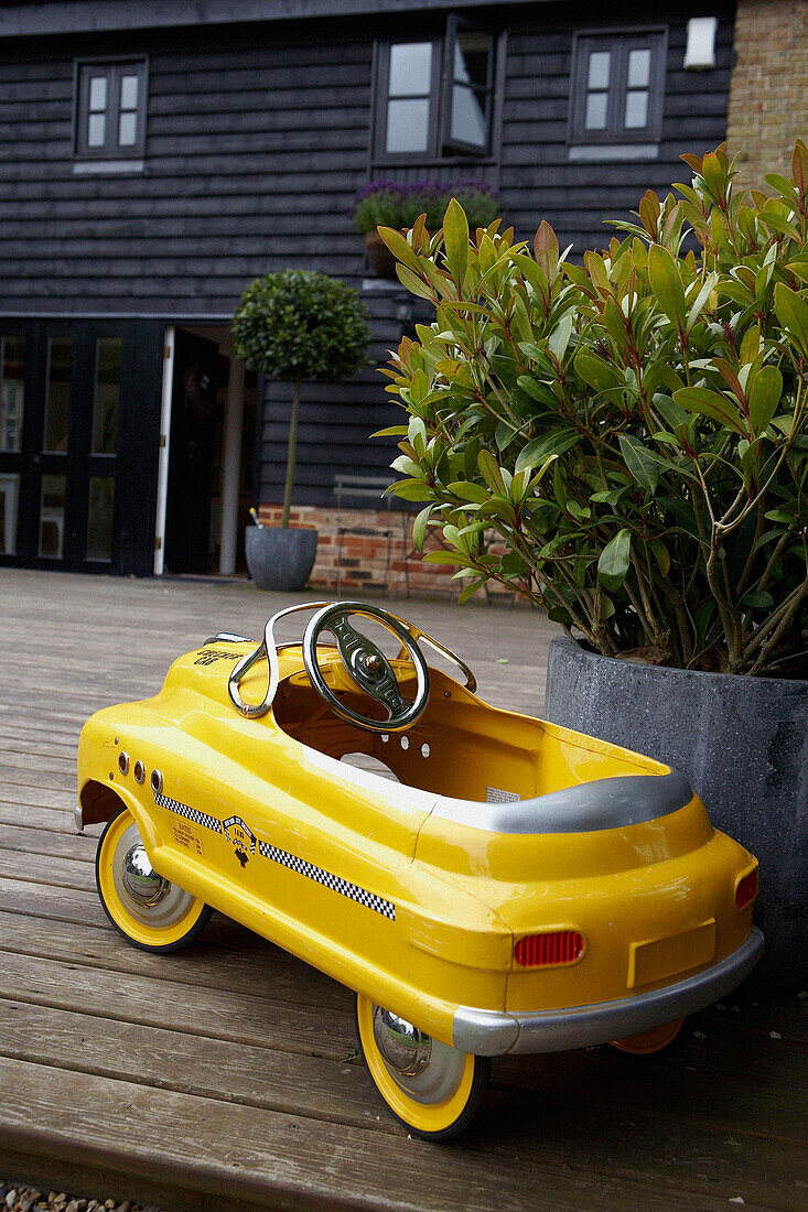 Leuchtend gelbes Kinderspielzeug-Tretauto-Taxi auf einer Gartenterrasse neben einer Topfpflanze