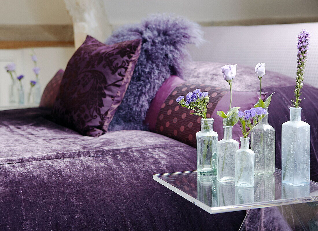 Medicine bottles with single stem flowers on table and purple bedspread with ostrich feather cushion renovated Cotswolds mill house England UK