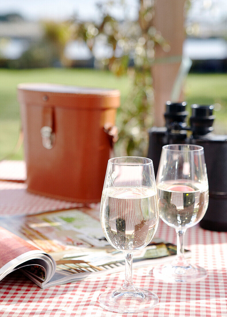 Weingläser und Fernglas auf einem Gartentisch mit aufgeschlagenem Magazin in einem Haus in Hampshire, England, UK
