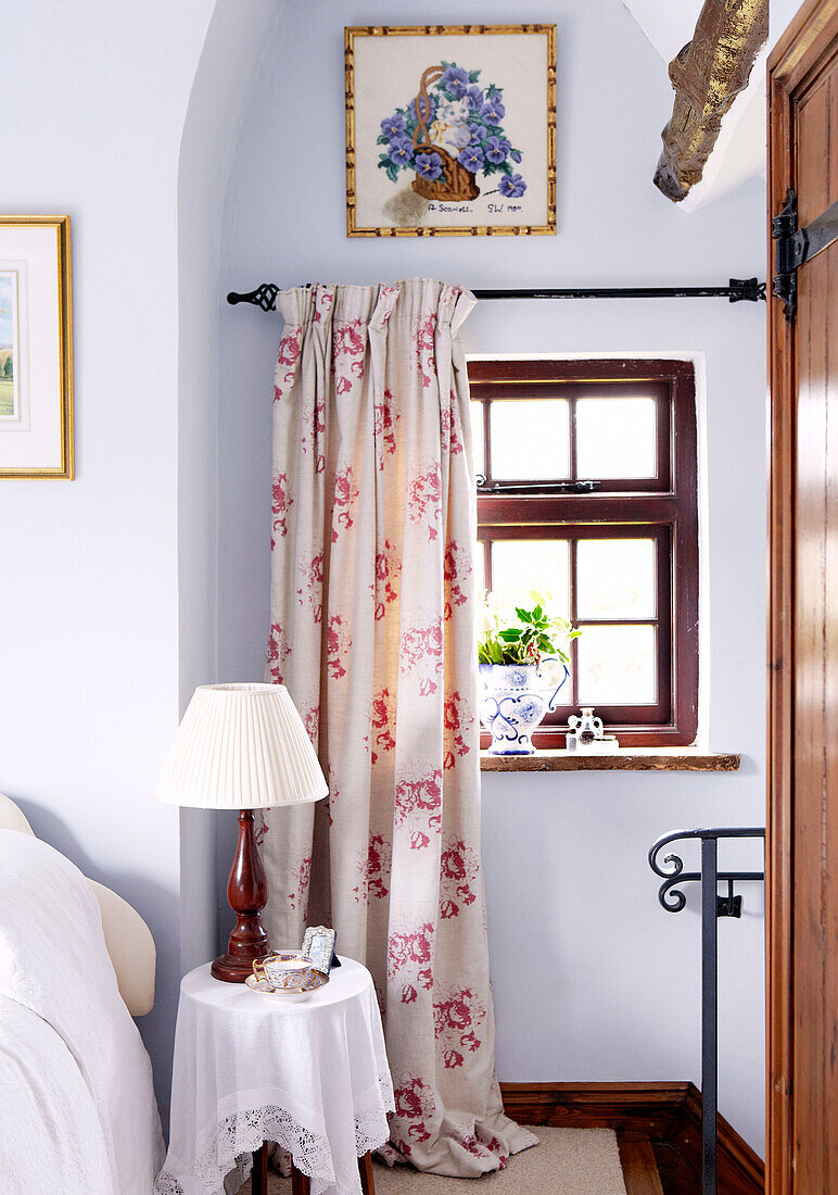 Lamp on side table below window with floral patterned curtain