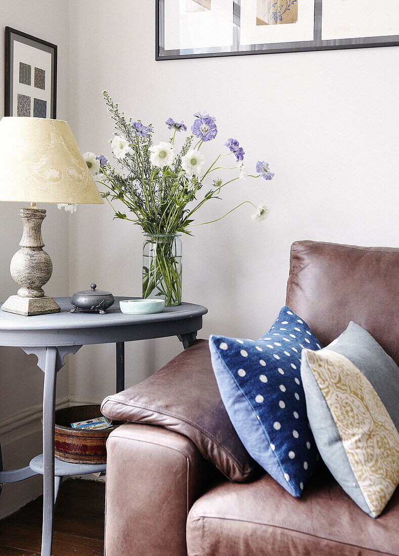Cut flowers on side table with spotted cushion on brown leather sofa in London home UK